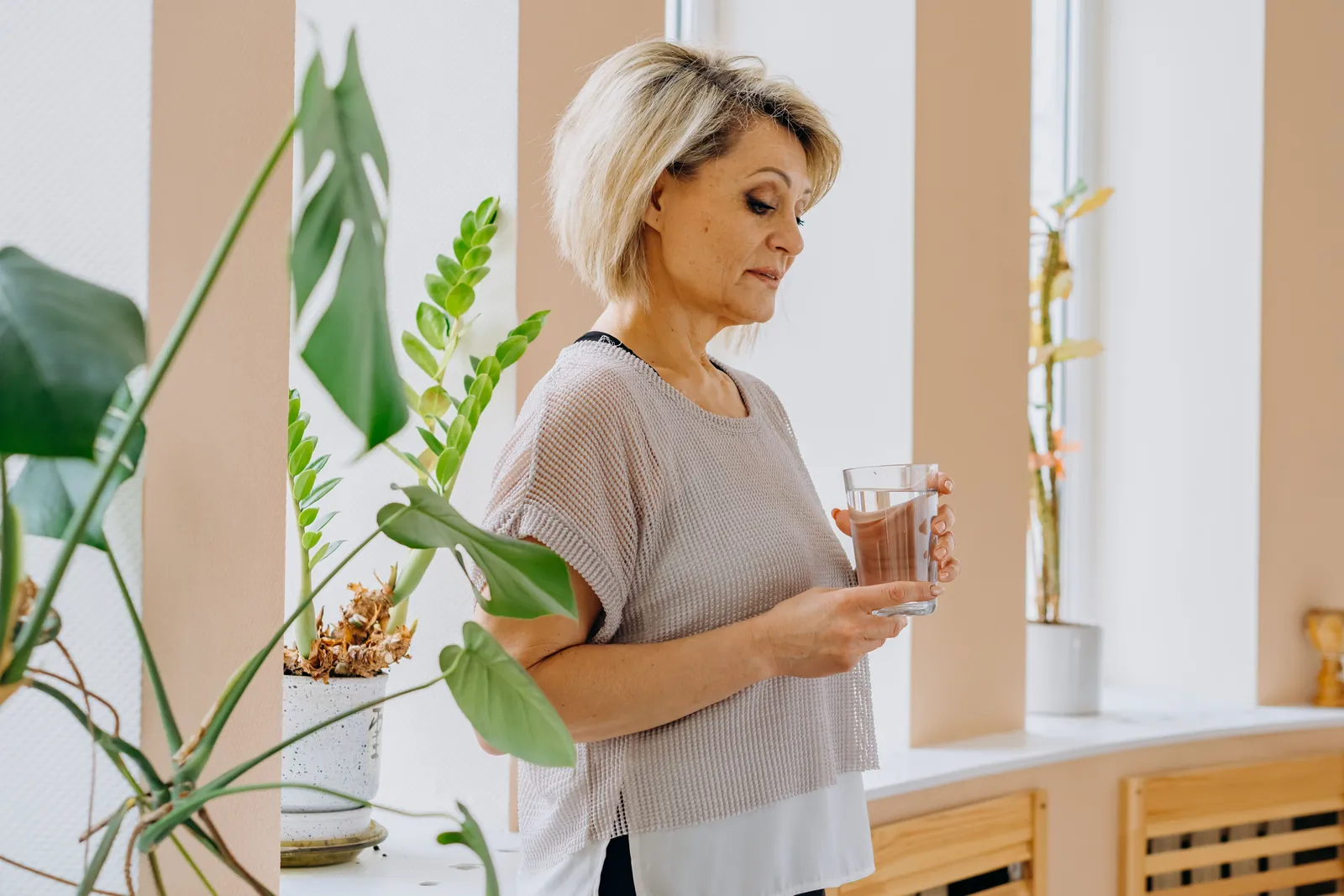 Een oudere vrouw met een glas water in een lichte woonkamer.